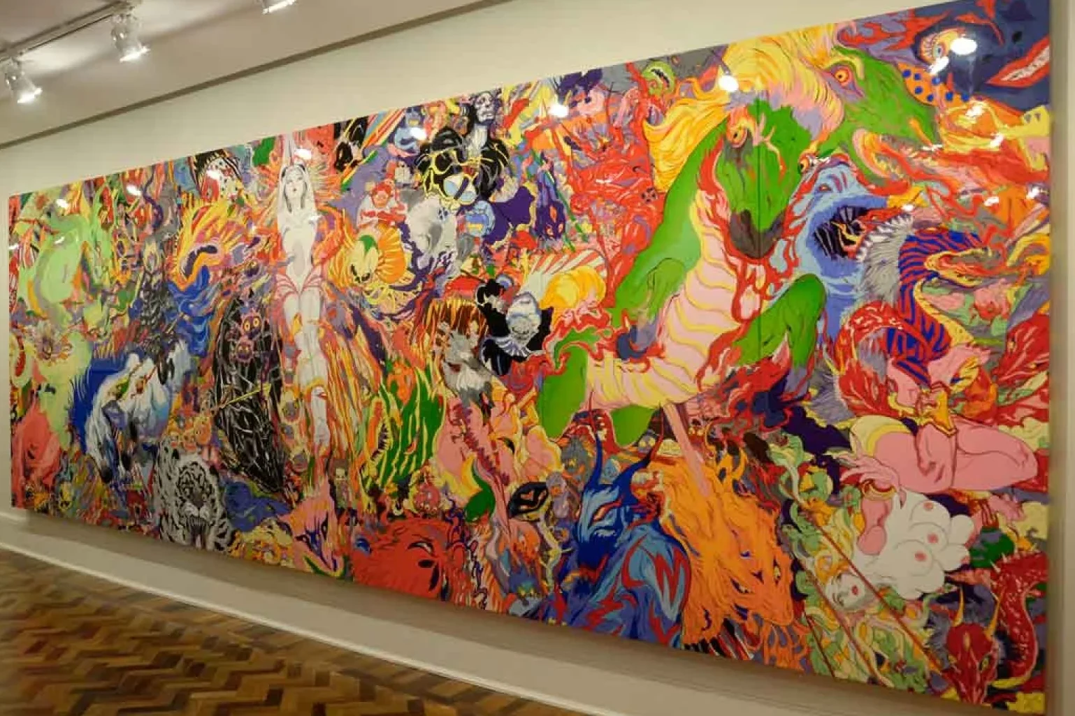 Fotografia de uma pintura monumental que ocupa quase toda a extensão de uma parede branca em uma galeria com piso de madeira em padrão espinha de peixe. A obra é um mural horizontal vibrante e densamente preenchido com figuras fantásticas e mitológicas em estilo que mistura anime e pop art. As cores são intensas e saturadas: amarelos, laranjas, verdes limão, azuis e rosas neon se entrelaçam em traços fluidos e orgânicos. À direita, destaca-se uma criatura verde que lembra um cavalo marinho gigante. No centro, figuras humanoides etéreas parecem flutuar em meio a explosões de cor e formas abstratas. A superfície da pintura é brilhante, refletindo sutilmente a iluminação dos spots no teto da galeria.